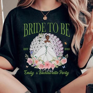 K&ouml;nnte beinhalten: Schwarzes T-Shirt mit dem Text "BRIDE TO BE" in Gr&uuml;n. Das Shirt zeigt eine Brautillustration in einem wei&szlig;en Kleid, umgeben von Blumen und Schmetterlingen. Der Text "Emily's Bachelorette Party" ist ebenfalls sichtbar.