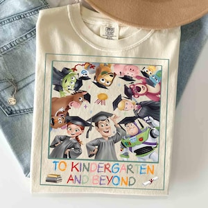 May include: Cream-colored t-shirt featuring a colorful graphic of Toy Story characters wearing graduation caps. The phrase "TO KINDERGARTEN AND BEYOND" is printed below the image in a playful font. The shirt is folded on a light blue denim surface.