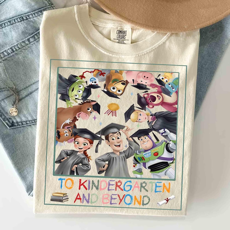 May include: Cream-colored t-shirt featuring a colorful graphic of Toy Story characters wearing graduation caps. The phrase "TO KINDERGARTEN AND BEYOND" is printed below the image in a playful font. The shirt is folded on a light blue denim surface.