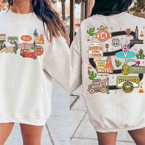 May include: Cream-colored sweatshirt featuring colorful graphics from the movie Cars. The front displays various signs and characters, while the back showcases a Route 66 design with cacti and other themed elements.