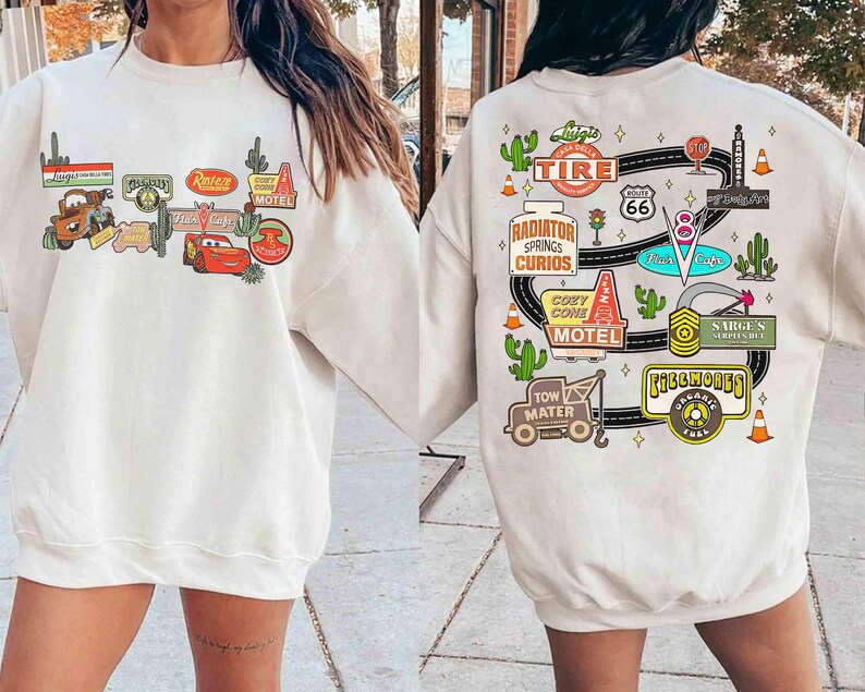 May include: Cream-colored sweatshirt featuring colorful graphics from the movie Cars. The front displays various signs and characters, while the back showcases a Route 66 design with cacti and other themed elements.