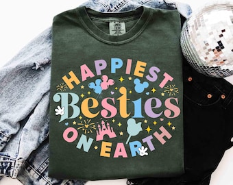 Könnte beinhalten: Dunkelgrünes T-Shirt mit dem Schriftzug "Happiest Besties on Earth" in einem farbenfrohen, skurrilen Design. Das Design umfasst Mickey Mouse-Silhouetten, Luftballons und ein Schloss. Das T-Shirt liegt gefaltet auf einer Jeansjacke und einer schwarzen Hose.