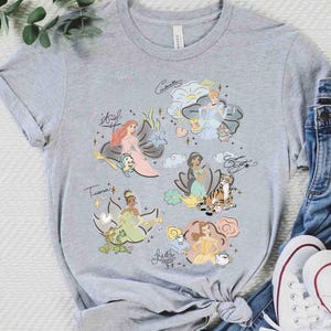 May include: A heather gray short-sleeve t-shirt featuring a graphic print of six animated princesses with their names written in cursive script. The design includes Ariel, Cinderella, Jasmine, Tiana, Belle, and various small animal companions.
