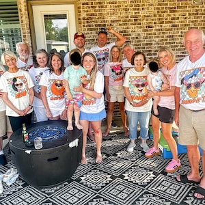 May include: A gathering of people wearing white t-shirts with cartoon dog designs and text. The shirts display sunglasses and the words "Mimi" and "Grandpa Tee". The group is outside near a fire pit.