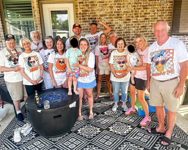 May include: A gathering of people wearing white t-shirts with cartoon dog designs and text. The shirts display sunglasses and the words "Mimi" and "Grandpa Tee". The group is outside near a fire pit.