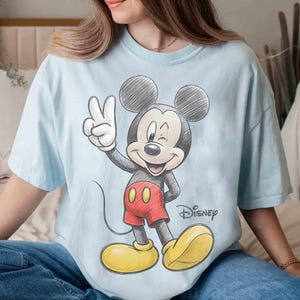 May include: Light blue t-shirt featuring a sketch-style Mickey Mouse graphic. Mickey is winking and giving a peace sign, wearing red shorts and yellow shoes. The Disney logo is printed below the character.