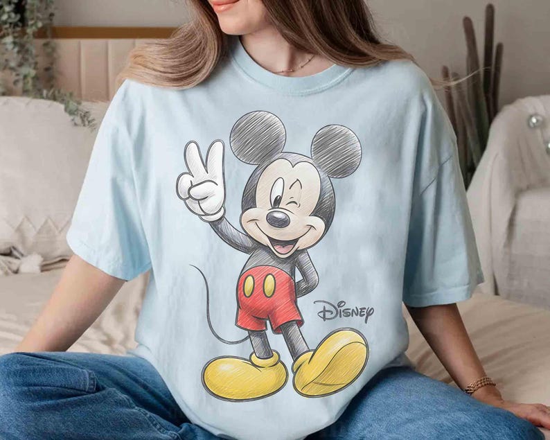 May include: Light blue t-shirt featuring a sketch-style Mickey Mouse graphic. Mickey is winking and giving a peace sign, wearing red shorts and yellow shoes. The Disney logo is printed below the character.