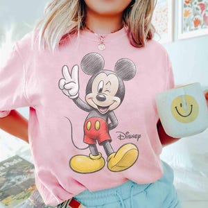 May include: Pink t-shirt featuring a sketch-style Mickey Mouse graphic. Mickey is giving a peace sign, wearing red shorts and yellow shoes. The word "Disney" is printed below the character.