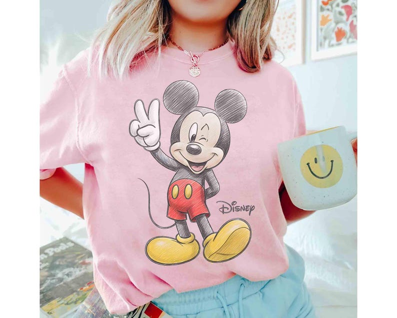 May include: Pink t-shirt featuring a sketch-style Mickey Mouse graphic. Mickey is giving a peace sign, wearing red shorts and yellow shoes. The word "Disney" is printed below the character.