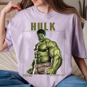 May include: A light purple t-shirt featuring a graphic of the Hulk. The Hulk is depicted in a muscular pose, with green skin and gray pants. The word "HULK" is printed above the image, with "SPIDER-MAN" and "BRAND NEW DAY" below.