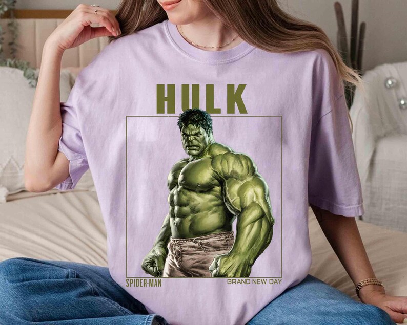 May include: A light purple t-shirt featuring a graphic of the Hulk. The Hulk is depicted in a muscular pose, with green skin and gray pants. The word "HULK" is printed above the image, with "SPIDER-MAN" and "BRAND NEW DAY" below.