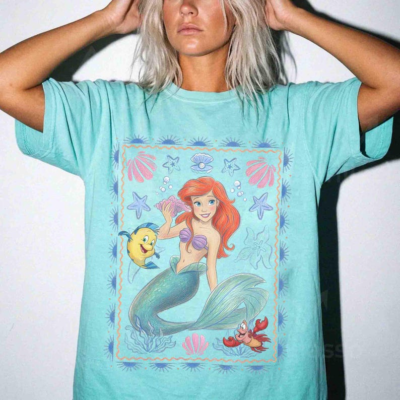 Op de afbeelding: Een licht turquoise t-shirt met een rechthoekige grafische print van een roodharige zeemeermin met een groene staart, een gele en blauwe vis en een kleine rode krab. Het ontwerp bevat schelpen, zeesterren en bubbels binnen een rand.