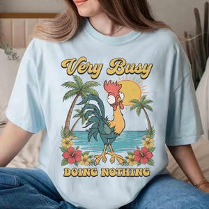 以下が含まれることがあります： 水色のTシャツ。漫画のニワトリのグラフィックがプリントされています。ニワトリはヤシの木、太陽、そして「Very Busy Doing Nothing」というテキストがあるビーチに立っています。デザインは黄色、オレンジ、緑色です。