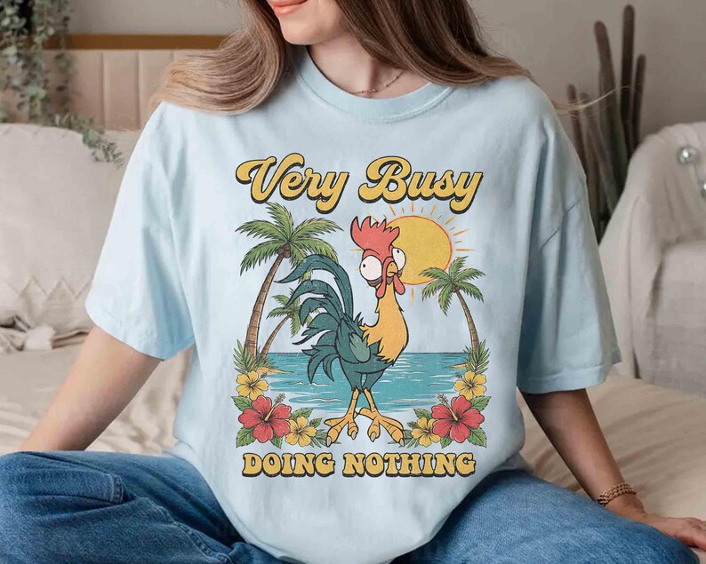 以下が含まれることがあります： 水色のTシャツ。漫画のニワトリのグラフィックがプリントされています。ニワトリはヤシの木、太陽、そして「Very Busy Doing Nothing」というテキストがあるビーチに立っています。デザインは黄色、オレンジ、緑色です。