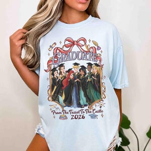 Pode incluir: Camiseta azul clara com um gr&aacute;fico de princesas de desenho animado em trajes de formatura. A palavra "GRADUATE" &eacute; exibida acima das princesas, com o texto "From The Tassel To The Castle 2026" abaixo. A camiseta tem mangas curtas.