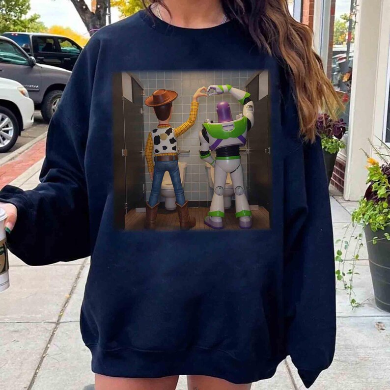May include: A dark navy blue crewneck sweatshirt featuring a graphic print of Woody and Buzz Lightyear standing in bathroom stalls, with their arms raised to form a heart shape together. The characters are depicted from behind in a tiled room.