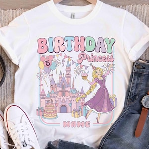 May include: White t-shirt with a colourful graphic design featuring the text "BIRTHDAY Princess" and a cartoon princess holding a cupcake. The design includes a castle, balloons, and the number "5".
