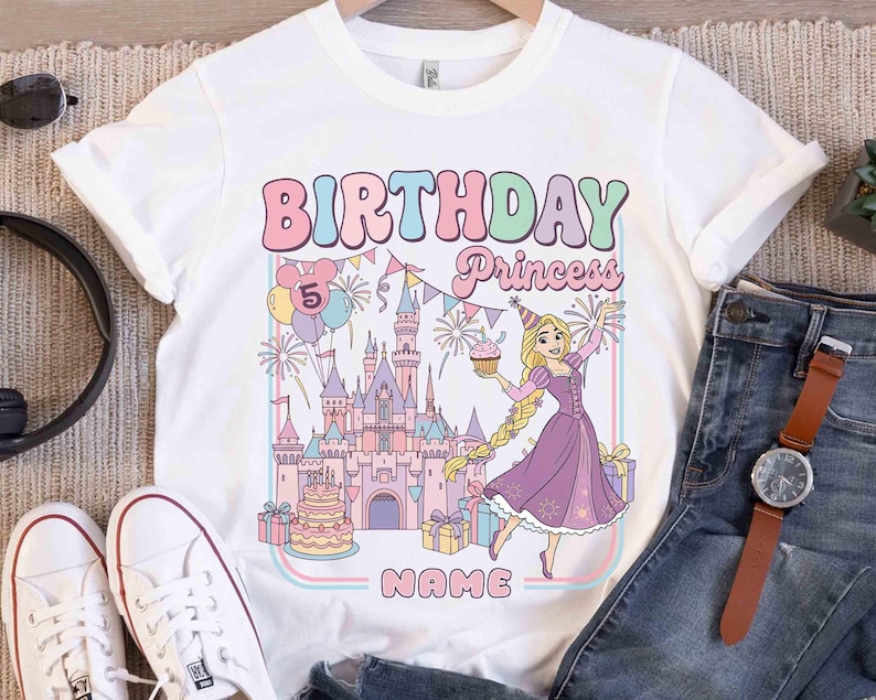 May include: White t-shirt with a colourful graphic design featuring the text "BIRTHDAY Princess" and a cartoon princess holding a cupcake. The design includes a castle, balloons, and the number "5".
