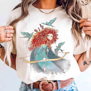 Puede incluir: Camiseta color crema con un gráfico de una mujer con cabello rizado rojo, sosteniendo un arco y flecha, con tres pájaros azules volando a su alrededor. El diseño tiene un efecto acuarela. La camiseta se combina con jeans azules y un cinturón marrón.