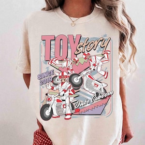 May include: Cream-coloured t-shirt with a graphic of Duke Caboom from Toy Story. The design includes the text "TOY STORY" and "Duke Caboom" with images of the character on a motorcycle. The shirt also has "SINCE 2019" printed on it.