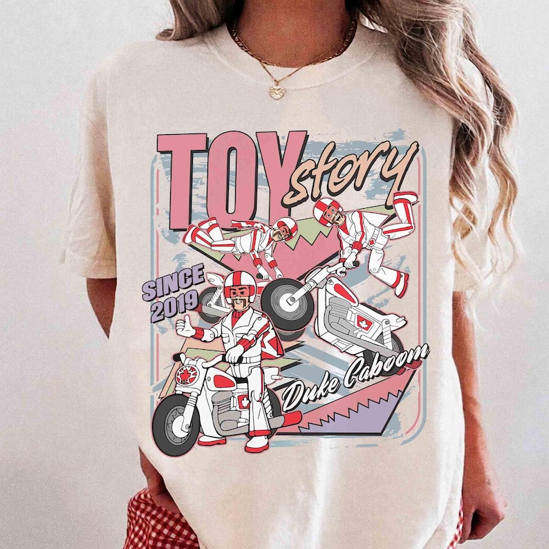 May include: Cream-coloured t-shirt with a graphic of Duke Caboom from Toy Story. The design includes the text "TOY STORY" and "Duke Caboom" with images of the character on a motorcycle. The shirt also has "SINCE 2019" printed on it.
