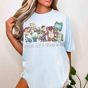 Op de afbeelding: Lichtblauw T-shirt met een afbeelding van personages uit de animatiefilm "Toy Story". Het ontwerp bevat Woody, Buzz Lightyear en andere personages, met de tekst "You've Got a Friend in Me" in bruin.
