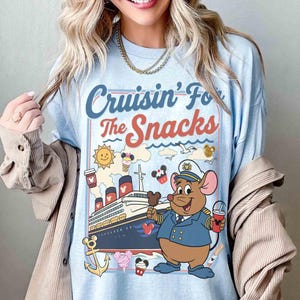 Pode incluir: Camiseta azul clara com o texto "Cruisin' For The Snacks" e uma ilustra&ccedil;&atilde;o de desenho animado de um navio de cruzeiro, um rato com uniforme de capit&atilde;o e v&aacute;rios &iacute;cones com tema de lanches. O design inclui um sol, sorvete e x&iacute;caras de caf&eacute;.