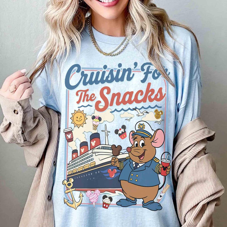 Pode incluir: Camiseta azul clara com o texto "Cruisin' For The Snacks" e uma ilustra&ccedil;&atilde;o de desenho animado de um navio de cruzeiro, um rato com uniforme de capit&atilde;o e v&aacute;rios &iacute;cones com tema de lanches. O design inclui um sol, sorvete e x&iacute;caras de caf&eacute;.