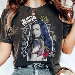 May include: Dark grey t-shirt with a graphic of a woman with blue hair, a crown, and the words "BEAUTY & BRAINS" in white. The design includes stars, hearts, and a red heart with a crown. The shirt is worn by a person.
