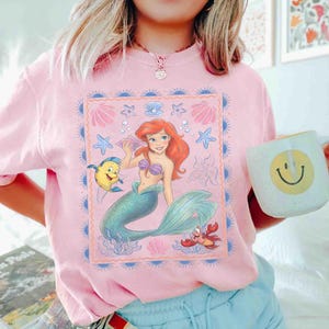 Op de afbeelding: Een lichtroze t-shirt met een rechthoekige grafische print van een roodharige zeemeermin met een groene staart, een gele vis en een rode krab. Het ontwerp is omringd door een blauwe rand met illustraties van schelpen en zeesterren.