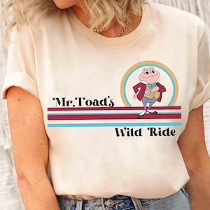 Könnte beinhalten: Cremefarbenes T-Shirt mit einem Cartoon-Bild von Mr. Toad in einem Kreis und dem Text "Mr. Toad's Wild Ride" darunter. Das Design umfasst bordeauxrote und türkisfarbene Streifen. Das Shirt hat kurze Ärmel.