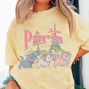May include: Yellow t-shirt featuring the word "Paris" in pink and blue, with the Eiffel Tower and three cartoon kittens. The word "Bonjour" is above the word "Paris".