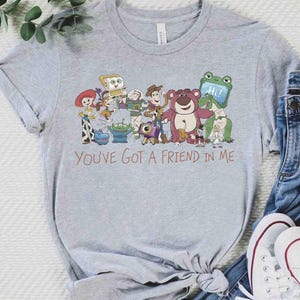 Op de afbeelding: Lichtgrijs t-shirt met een kleurrijke afbeelding van personages uit de animatiefilm "Toy Story". De zin "You've Got a Friend in Me" staat in bruin schrift eronder. Het shirt wordt gecombineerd met een blauwe jeans en witte sneakers.