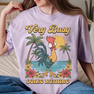 以下が含まれることがあります： 「Very Busy Doing Nothing」の文字が黄色と黒でプリントされた薄紫色のTシャツ。漫画のニワトリ、ヤシの木、太陽、熱帯の花が描かれたグラフィックデザインです。