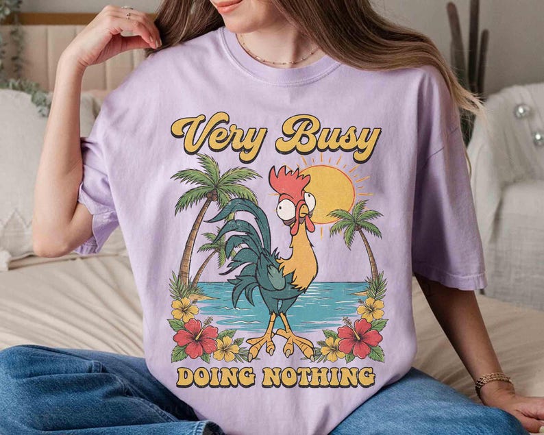以下が含まれることがあります： 「Very Busy Doing Nothing」の文字が黄色と黒でプリントされた薄紫色のTシャツ。漫画のニワトリ、ヤシの木、太陽、熱帯の花が描かれたグラフィックデザインです。