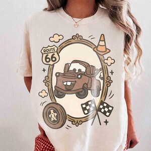 May include: Cream-colored t-shirt featuring a cartoon image of a brown tow truck inside a decorative frame. The design includes a Route 66 sign, a traffic cone, a tire, clouds, and checkered flags. The shirt has short sleeves.