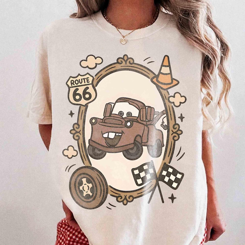 May include: Cream-colored t-shirt featuring a cartoon image of a brown tow truck inside a decorative frame. The design includes a Route 66 sign, a traffic cone, a tire, clouds, and checkered flags. The shirt has short sleeves.