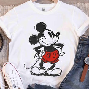 May include: White t-shirt featuring a black and red graphic of Mickey Mouse. The cartoon character is in a sketched style, with his hands on his hips and a smiling expression. The shirt is displayed with jeans and accessories.