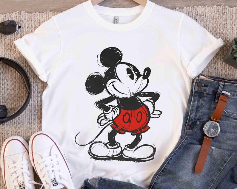May include: White t-shirt featuring a black and red graphic of Mickey Mouse. The cartoon character is in a sketched style, with his hands on his hips and a smiling expression. The shirt is displayed with jeans and accessories.