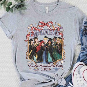 Pode incluir: Camiseta cinza claro com um gr&aacute;fico colorido de princesas da Disney em trajes de formatura. A palavra "GRADUATE" &eacute; exibida acima das princesas, com o texto "From The Tassel To The Castle 2026" abaixo.