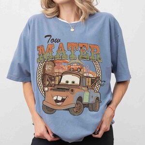 K&ouml;nnte beinhalten: Ein staubblaues Kurzarm-T-Shirt mit einem Grafikdruck der Figur Tow Mater aus dem Film Cars. Das Design enth&auml;lt den Schriftzug Tow Mater, einen kreisf&ouml;rmigen Rahmen, einen Wasserturm und ein Radiator Springs Racers-Schild in Orange und Gr&uuml;n.