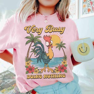 以下が含まれることがあります： 「Very Busy Doing Nothing」の文字が入ったピンク色のTシャツ。ヤシの木、太陽、花のあるビーチにいる漫画のニワトリのグラフィック。笑顔の白いマグカップが手に持たれています。