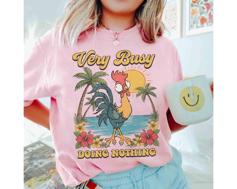 以下が含まれることがあります： 「Very Busy Doing Nothing」の文字が入ったピンク色のTシャツ。ヤシの木、太陽、花のあるビーチにいる漫画のニワトリのグラフィック。笑顔の白いマグカップが手に持たれています。