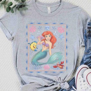 Op de afbeelding: Een grijs gem&ecirc;leerd t-shirt met korte mouwen en een rechthoekige grafische print van een zeemeermin met rood haar en een groenblauwe staart, een gele vis en een rode krab. Het ontwerp bevat roze schelpen, blauwe zeesterren en een zonnestraalrand.