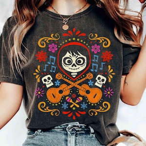 May include: Dark gray t-shirt featuring a colorful graphic design inspired by the movie Coco. The design includes a skull, guitars, musical notes, and floral elements in red, orange, yellow, and blue.