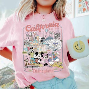Puede incluir: Camiseta rosa claro con un dise&ntilde;o de Disneyland que presenta a Mickey Mouse, Minnie Mouse, Donald Duck y otros personajes. El dise&ntilde;o incluye el texto "California Est. 1955" y "Disneyland". En la imagen se sostiene una taza blanca con una carita sonriente amarilla.