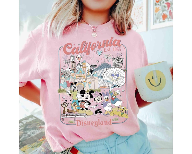 Puede incluir: Camiseta rosa claro con un dise&ntilde;o de Disneyland que presenta a Mickey Mouse, Minnie Mouse, Donald Duck y otros personajes. El dise&ntilde;o incluye el texto "California Est. 1955" y "Disneyland". En la imagen se sostiene una taza blanca con una carita sonriente amarilla.