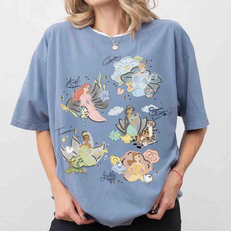 May include: A dusty blue t-shirt featuring a graphic print of five animated princesses, including Ariel, Cinderella, Jasmine, Tiana, and Belle. Each character is accompanied by their signature, small icons, and decorative floral elements in soft pastel colors.