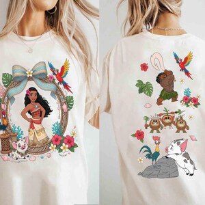 May include: A cream-colored t-shirt featuring a front graphic of a character in a patterned frame with a pig, a rooster, and a blue bow, and a back graphic showing a male character with a hook, three small coconut creatures, a rooster, and a pig.