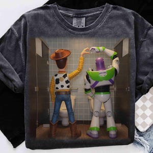 May include: A charcoal gray mineral-washed t-shirt featuring a graphic print of Woody and Buzz Lightyear standing in bathroom stalls, forming a heart shape with their hands. The characters are shown from behind, wearing their signature outfits.